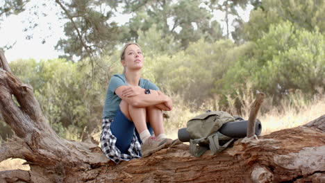 Young-Caucasian-woman-sits-on-a-fallen-tree,-looking-thoughtful