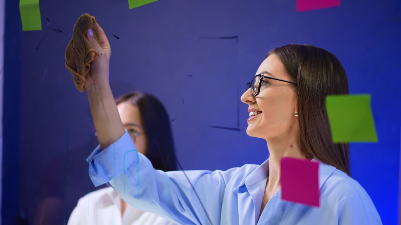 Positive smiling Caucasian brunette in glasses cleans the glass board. Colorful stickers with ideas are around on the glass. Close up.