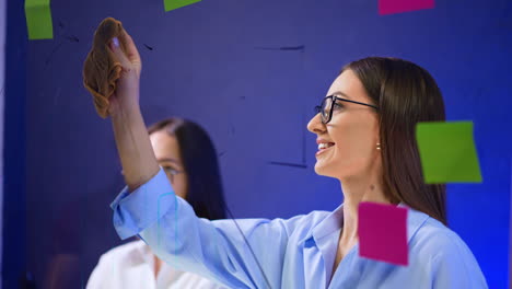positive smiling caucasian brunette in glasses cleans the glass board. colorful stickers with ideas are around on the glass. close up.