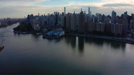 La-Cámara-Aérea-épica-Se-Inclina-Hacia-Arriba-Para-Revelar-El-Horizonte-De-La-Ciudad-De-Nueva-York-Con-Un-Bonito-Reflejo-En-El-Río-Este-Al-Atardecer-La-Inclinación-Sigue-Subiendo-Hacia-El-Cielo-Azul