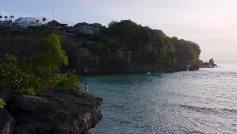 zoomed out shot of young girl clad in bikini standing on clifftop at beachside and enjoying beautiful landscape