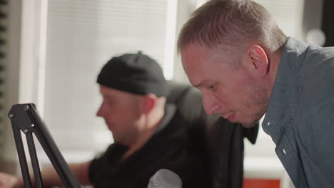 a close-up of a man in a studio leaning over and singing into a microphone, while another man, likely a producer, dressed in black, is seated and focused on the computer screen