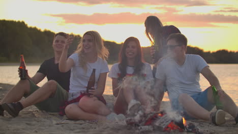 the young people are sitting in around bonfire on the river coast. they are laughing and enjoying beer at sunset in summer evening.