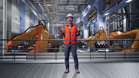 full body of asian male engineer with safety helmet smiling to camera and saying wow while standing with industrial robotic arms working