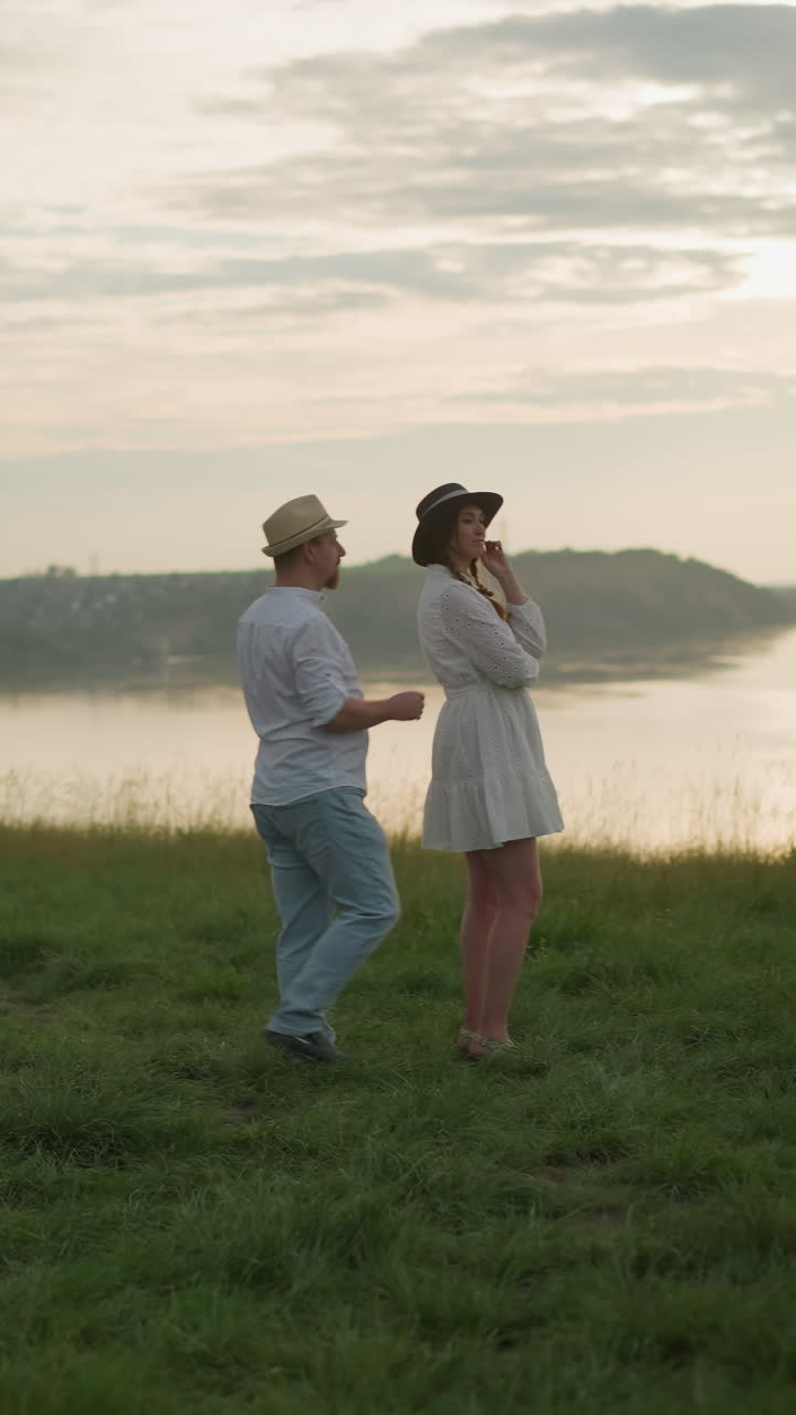 모자,  셔츠, 청바지를 입은 남자가  드레스를 입은 여자를 향해 걸어가는 조용한 호수를 내려다보는 잔디로 인 언덕 위에 서 있는 조용한 장면.