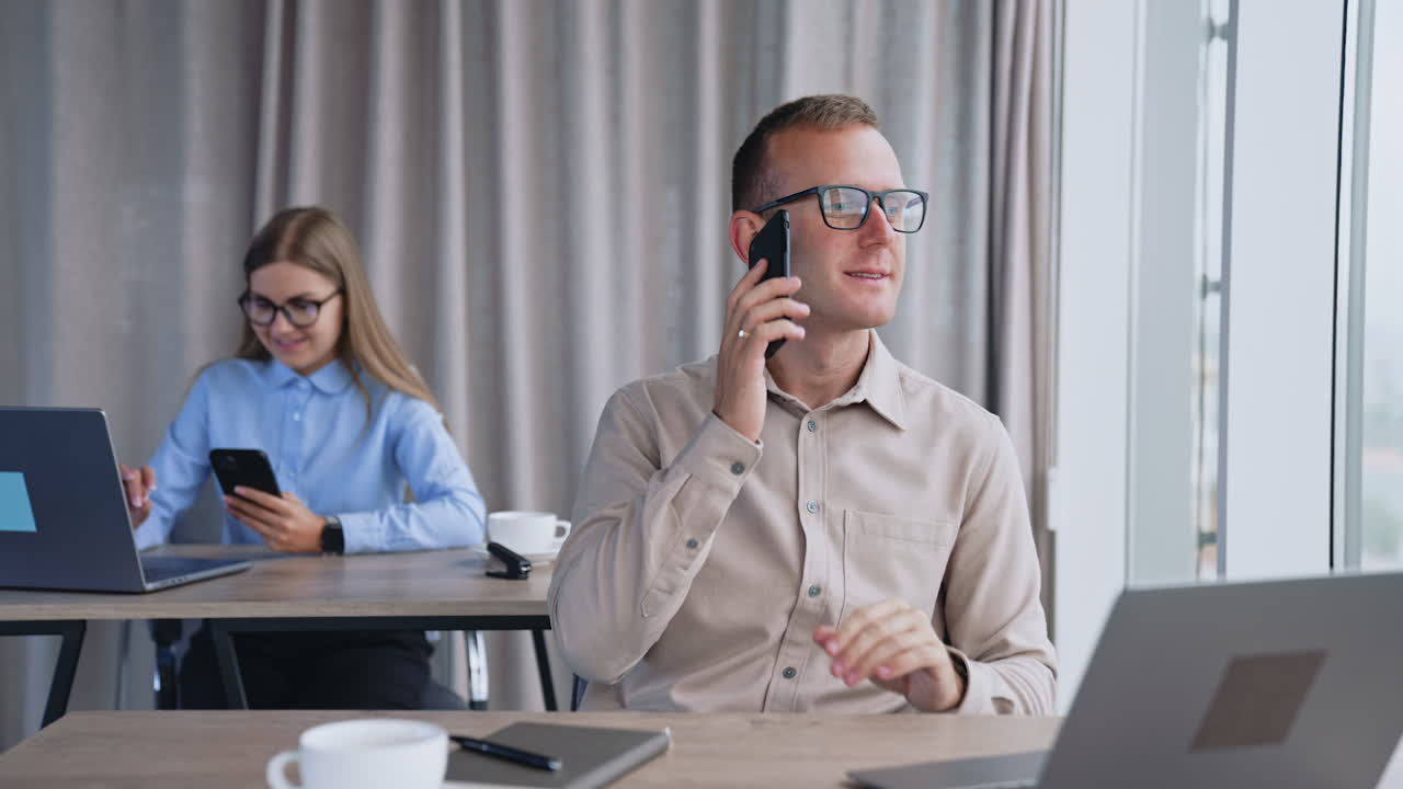 Having a phone call in office. Man speaks on his phone smiling. Close up. Lady at backdrop uses phone and laptop at once.