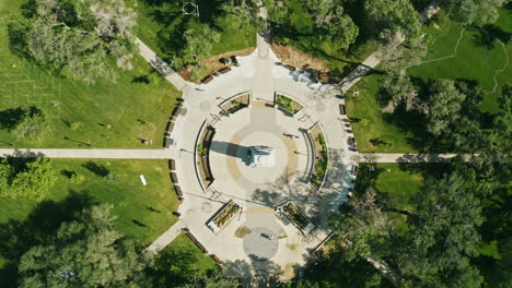 aerial cenital shots over a park with a square and a monument in its center with symmetrical paths and a person walking