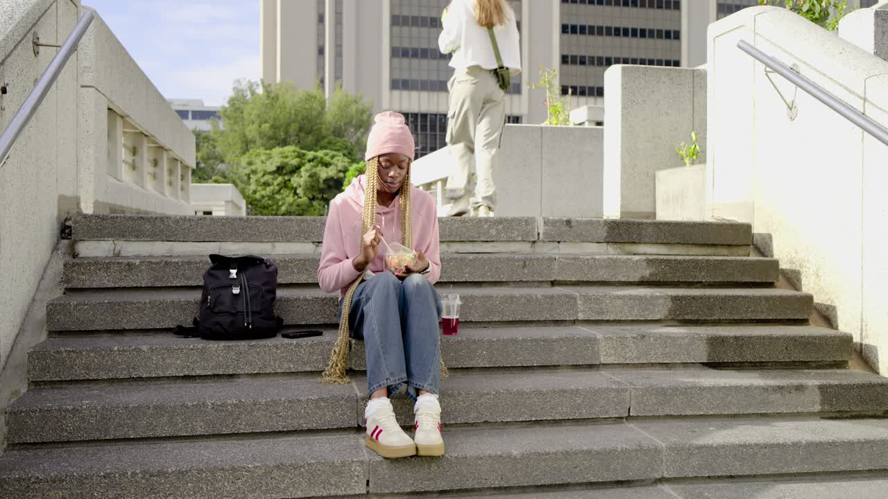 Sitting woman eating takeout on stairs with backpack while passing woman carrying phone and folder