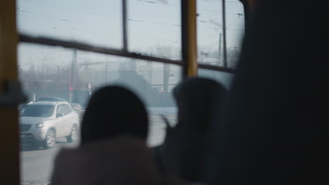 inside moving bus, passengers in winter coats watch opposite tram glide past outside, view through windows with soft blur on glass, muted city light, motion on snowy street during urban commute