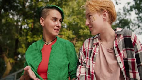 Close-up-shot-of-a-girl-with-short-green-hair-in-a-green-shirt-and-a-blonde-girl-with-short-hair-in-a-checkered-shirt-talking-on-their-date-in-the-park.-Two-lesbians-in-the-park-on-a-date