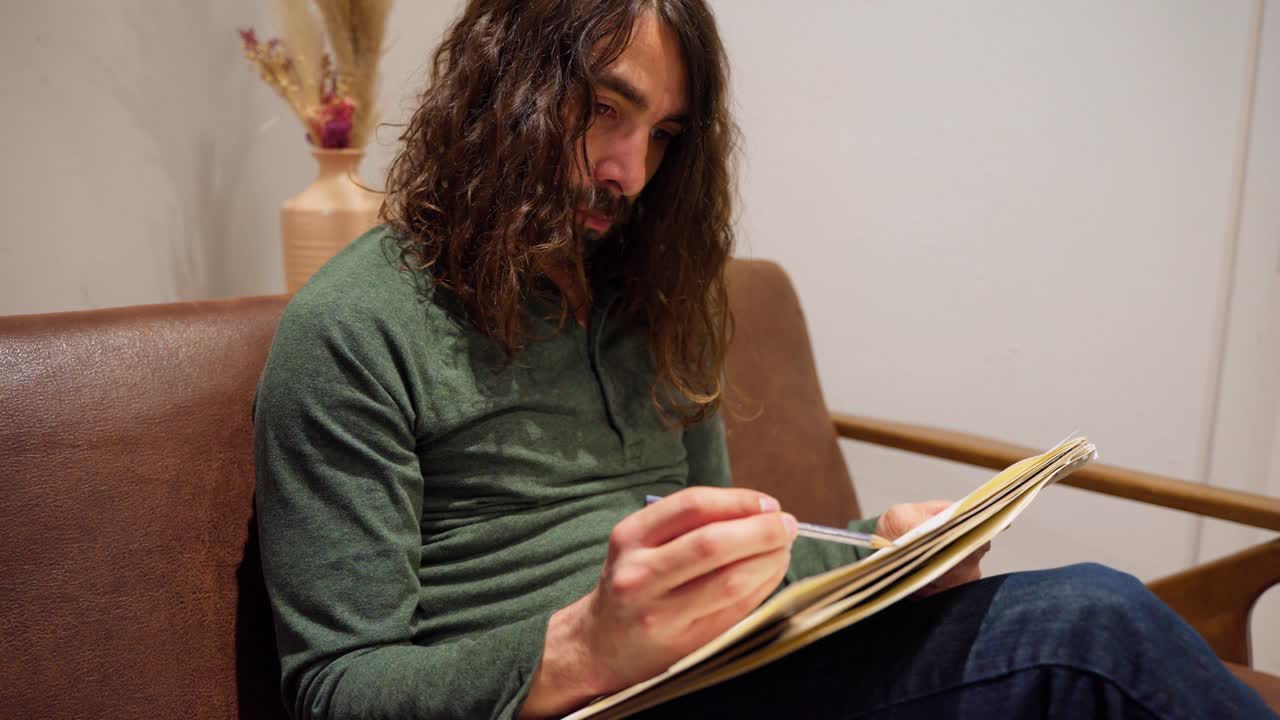 artista masculino étnico con cabello largo y barba en camisa verde sentado en un sofá marrón dibujando
