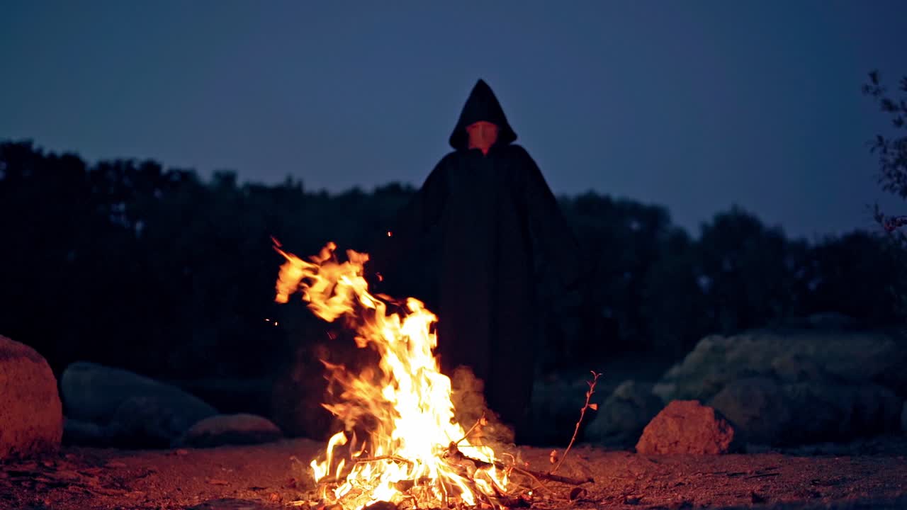 Dark wizard in hood behind the burning fire. Scary death with outstretched arms doing shamanism. Black ghost performing ritual near the fire. Horror concept.