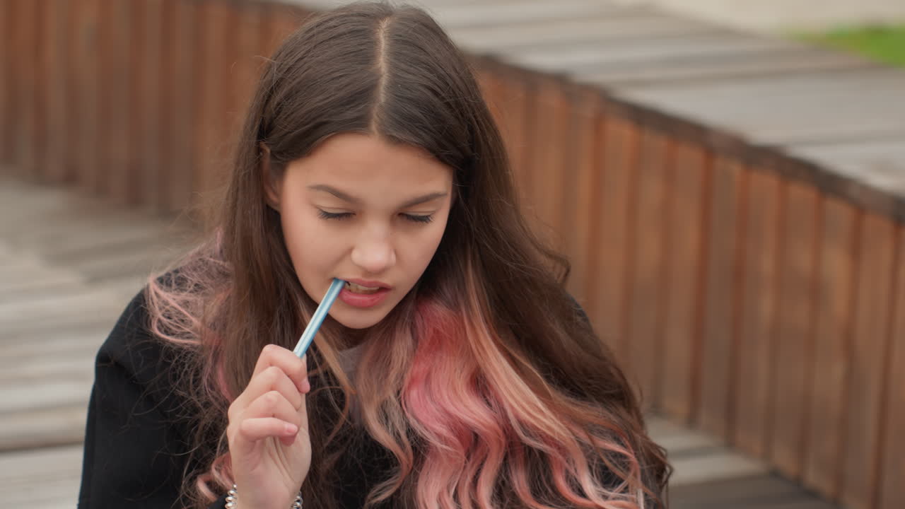 Studente bianco concentrato che studia su una panchina, rosicchiando la penna mentre prende appunti, campus con passerella in legno, concentrazione introspettiva, toni autunnali e routine di studio disciplinata per adolescenti