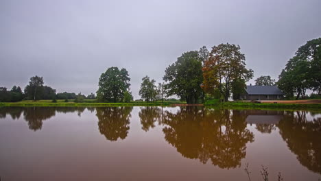 Timelapse-View-Over-Brown-Lake-With-Overcast-Clouds-Rolling-Past-And-Rustic-Cabin-In-Background