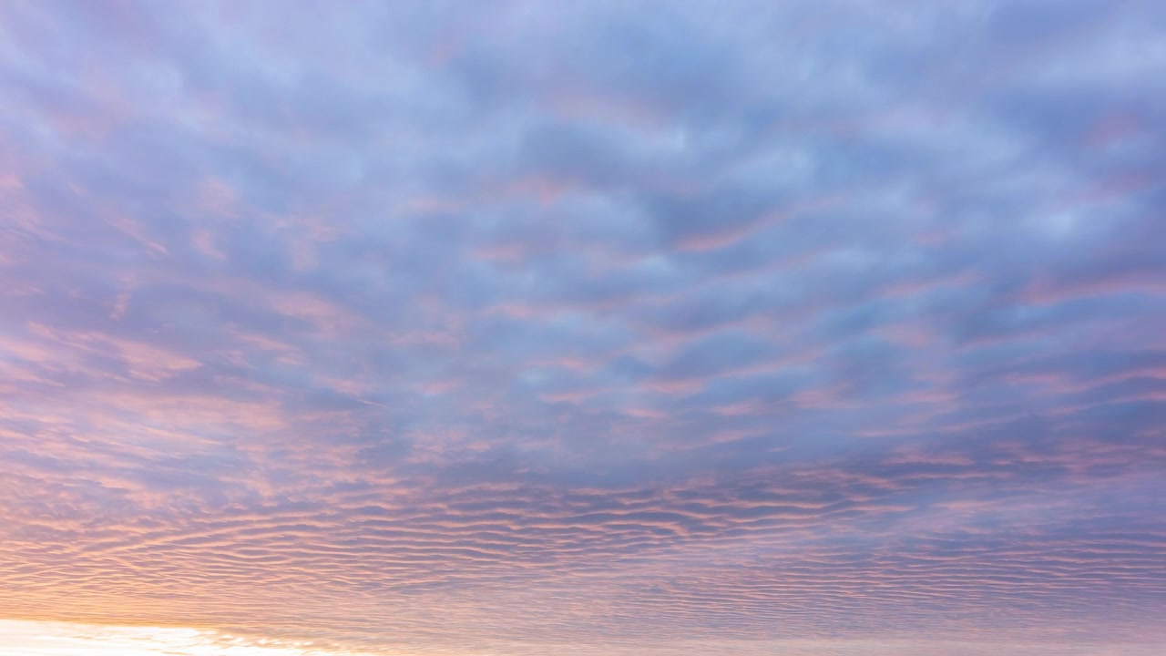 Wide view of a sunset sky with soft layered clouds over a tranquil horizon, warm tones of orange and pink illuminate the atmosphere creating a serene evening