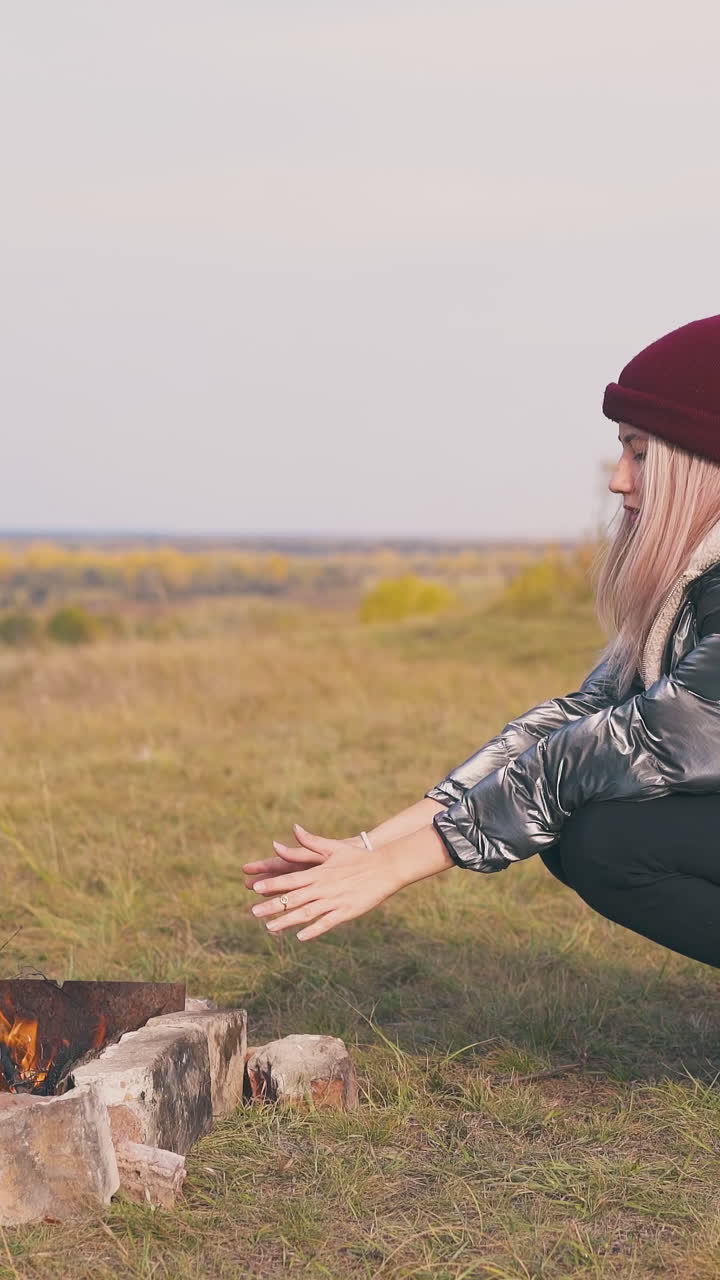 pretty young woman heats palms at burning bonfire on river bank meadow against clear sky in morning