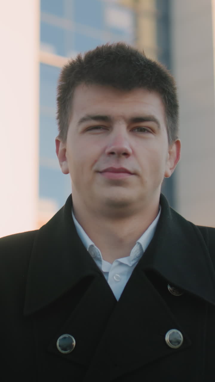 hombre con abrigo negro se encuentra al aire libre con una sonrisa sutil, que irradia confianza y equilibrio, el fondo borroso con edificios modernos destaca el entorno profesional urbano