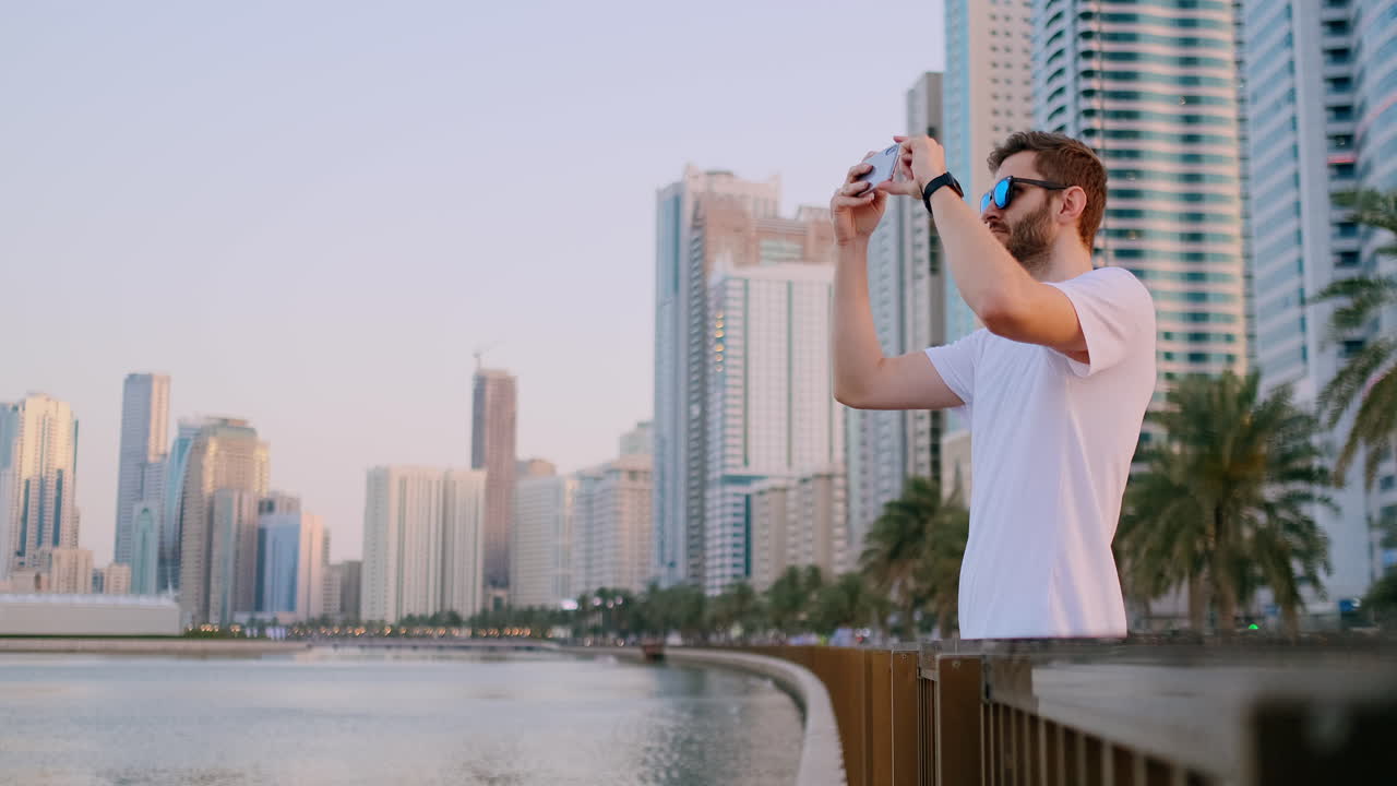 A young man in a white t-shirt standing on the waterfront against the background of the modern city takes photos and videos on the phone for social networks is live
