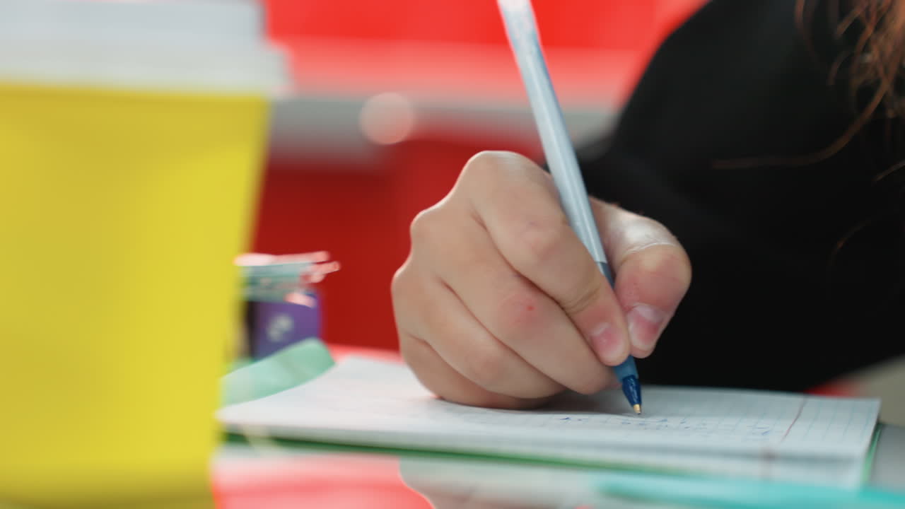 Studerende skriver med blå kuglepen i notesbog, nærbillede af hånd nær gul kop, farverige kontorartikler, lys klasseværelsesbaggrund, afslappet kropsholdning, kreativ notetagning, forberedelse til gruppearbejde, studiepause