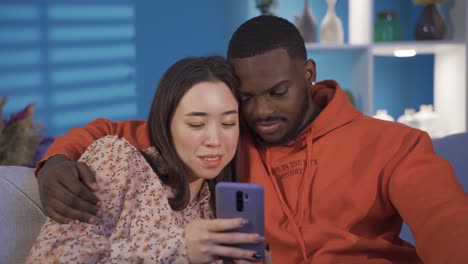 happy multiracial couple sitting on sofa at home chatting, looking at phone and laughing.