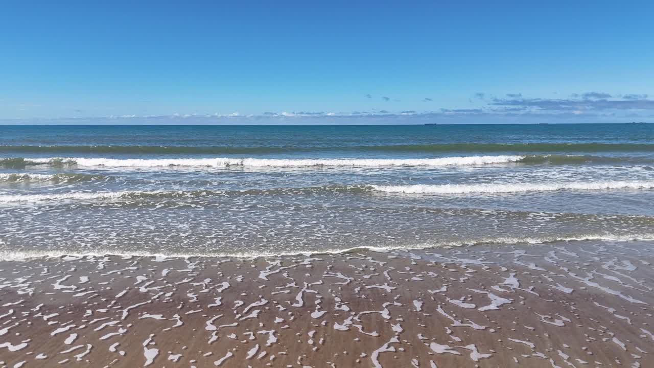 Calm ocean waves roll onto sandy beach under clear blue sky, steady camera, natural daylight