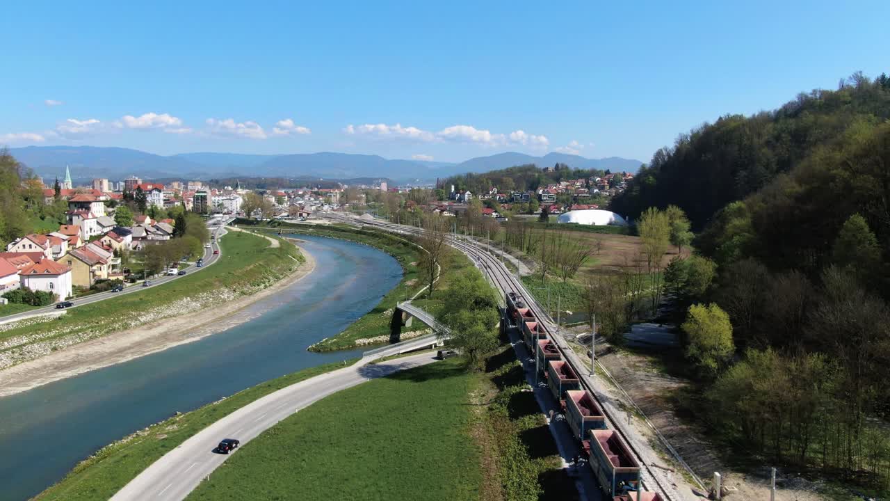 Drone follows a goods train that runs along a river heading to a city. Drone shot