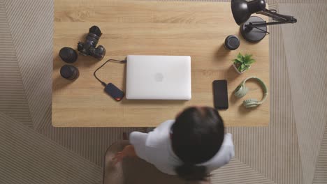 top view of a woman editor walking into the workspace sitting down and using a laptop next to the camera editing photo of a woman at home