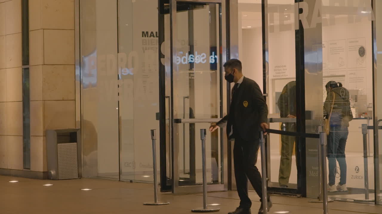 Guardia di sicurezza con mascherina che posiziona paletti delimitatori all'esterno di un edificio moderno di notte.