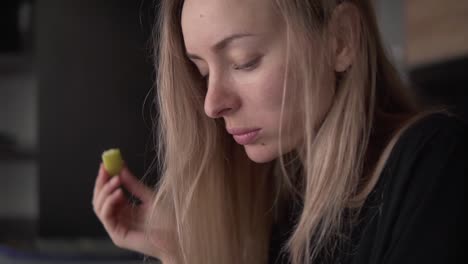 blonde young woman standing at the kitchen eating apple indoors at home