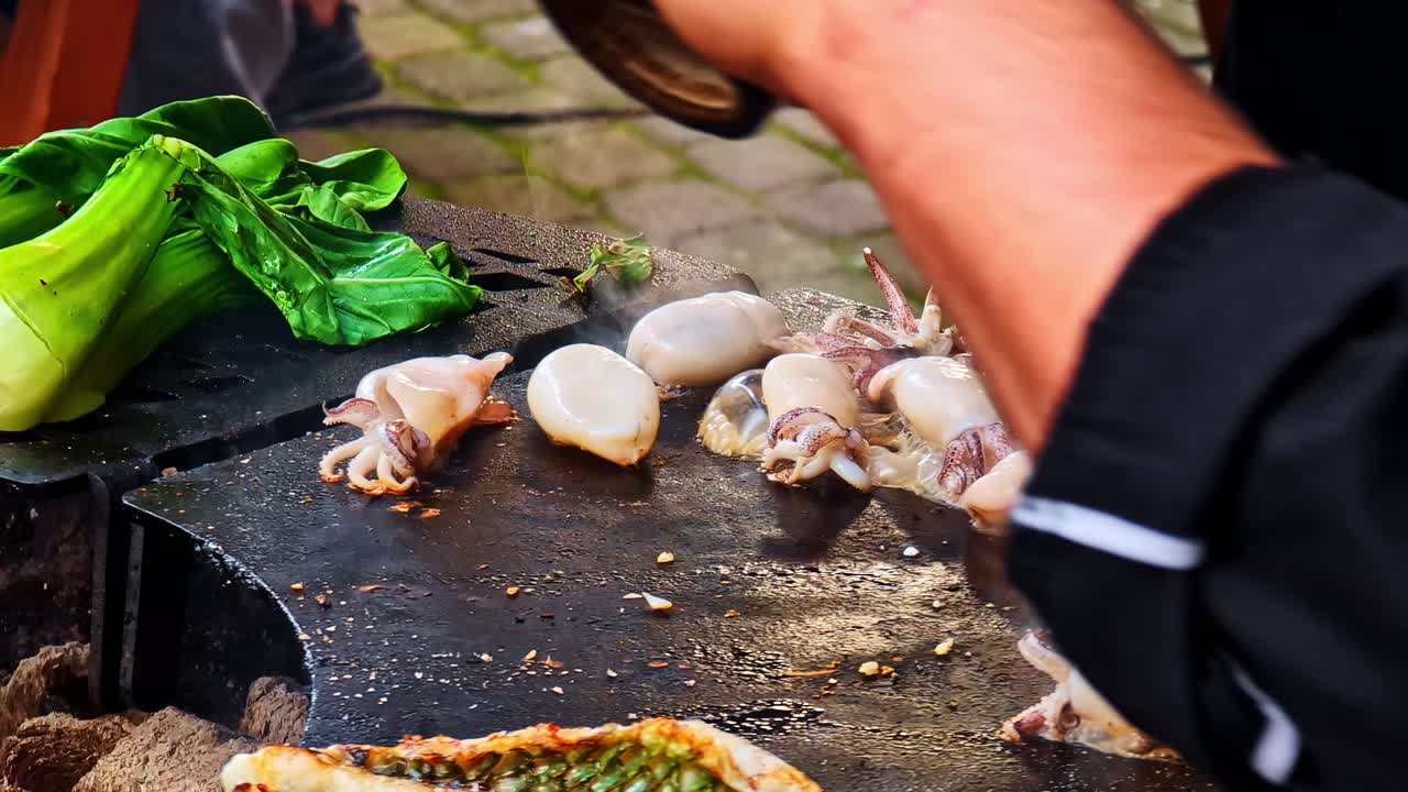 Grilled squid and fish on open charcoal grill with tongs flipping seafood for cooking