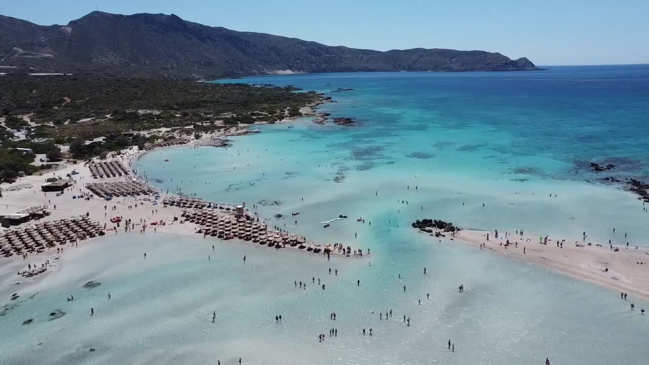 Breathtaking aerial footage of Elafonissi Beach, Santorini. Turquoise waters meet sandy shores under a sunny sky, with sunbathers and umbrellas dotting the shoreline.