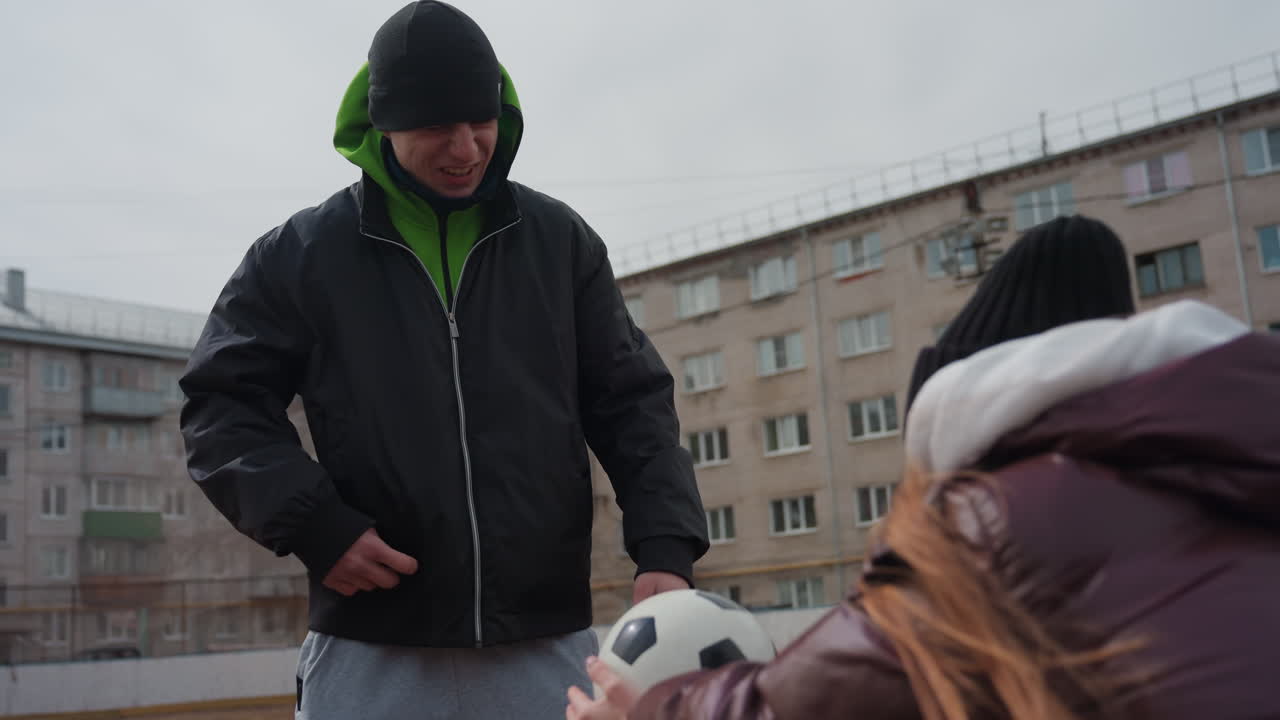 Белый тренер мужского пола передает футбольный мяч на городской площадке, давая советы ребенку, холодный пасмурный день, многоквартирные дома на заднем плане, куртка с капюшоном и пуховик, дружелюбное общение, момент наставничества