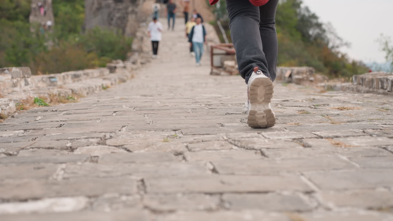 Primi piani di passi su ciottoli irregolari, un passo fermo che sale un sentiero consumato, con un'angolazione bassa della fotocamera che cattura i dettagli della suola e del battistrada. Altri camminatori appaiono sullo sfondo, con un ritmo in salita e un'atmosfera storica.