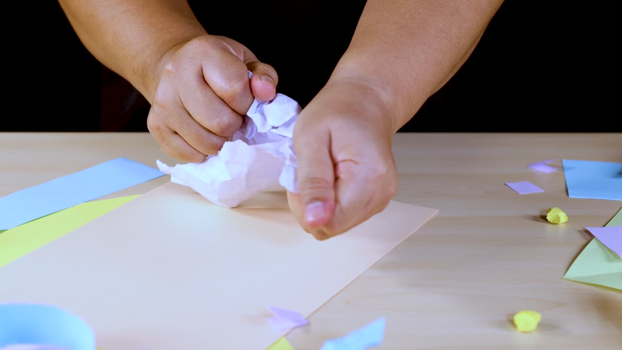 Person tears and crumples white paper on a pastel craft table under bright studio lighting