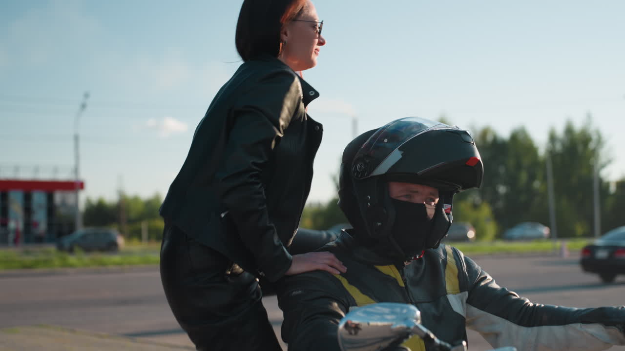 Motorcycle rider in helmet and leather jacket opens visor while woman in leather climbs onto bike smiling, preparing for ride off under clear sky and urban street setting