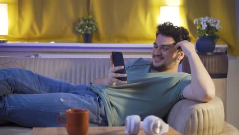 hombre acostado en el sofá por la noche enviando mensajes de texto por teléfono. feliz y de buen humor.