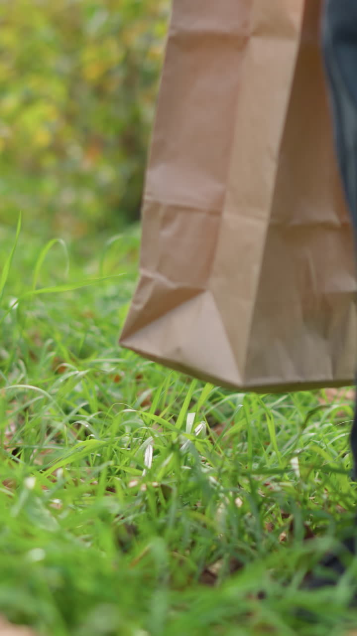 vista de la pierna de un niño con pantalones vaqueros oscuros y zapatillas negras caminando a través de la vegetación vibrante, sosteniendo una bolsa de papel marrón, rodeado de plantas exuberantes y follaje en un entorno natural al aire libre