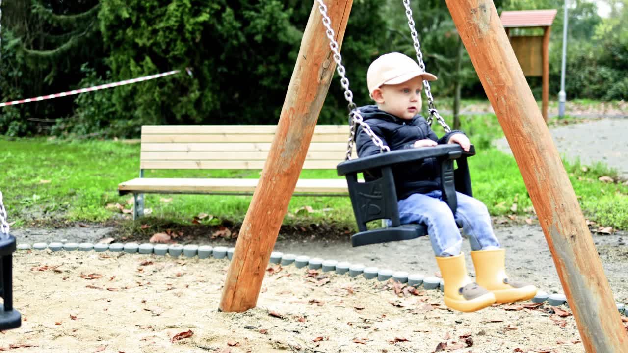 Cheerful 2 year old boy swings on a swing on a playground. Toddler swings on a swing in a city park. Child sits on a children's swing. Children's attractions