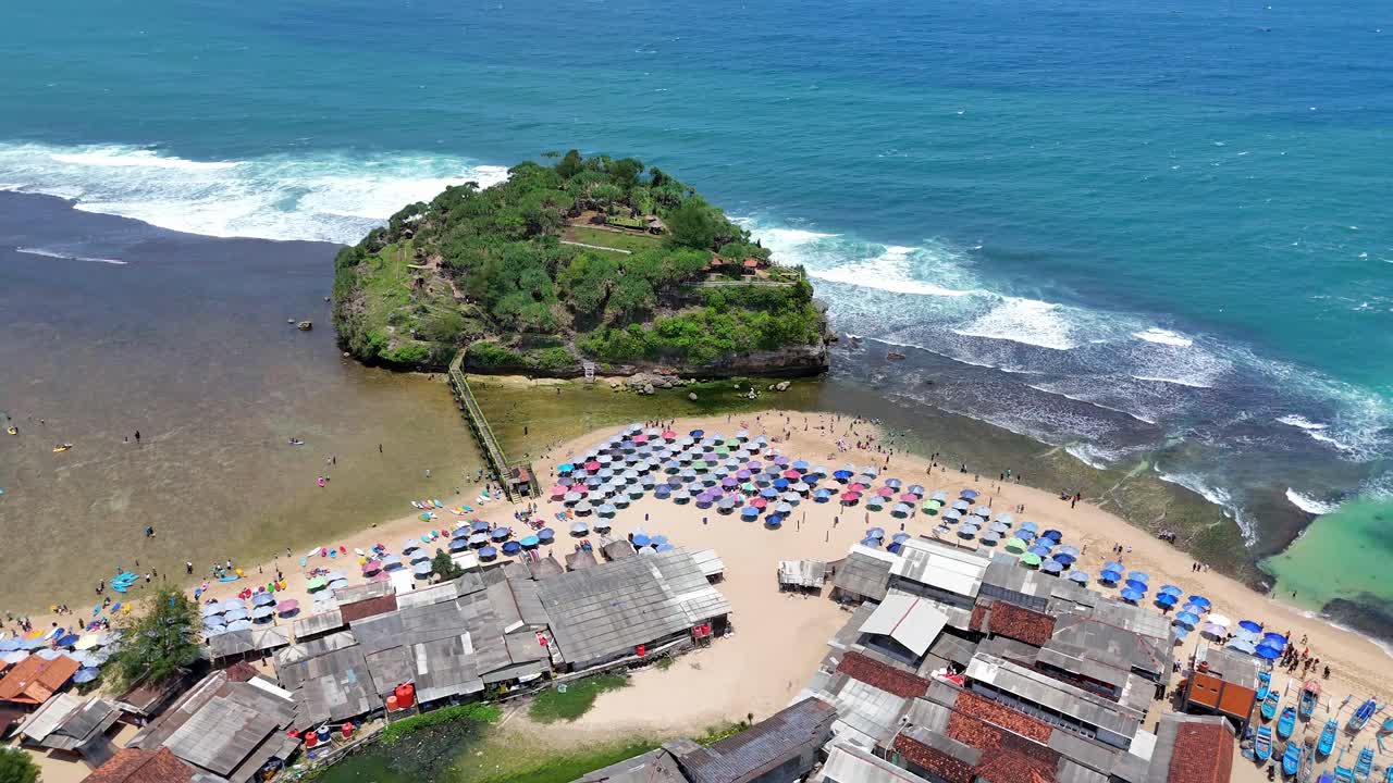 Splendido filmato aereo di una spiaggia tropicale con file di ombrelloni colorati, un ponte di legno che collega a una lussureggiante isola verde e onde blu cristalline che si infrangono sulla costa rocciosa