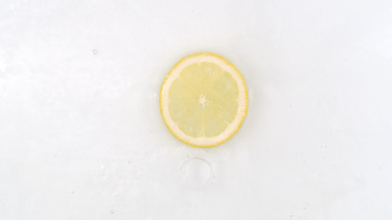 On a white background a splash of water falls on a slice of lemon in slow motion.