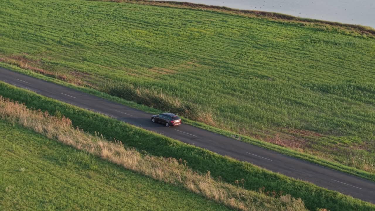 A brown car drives along a quiet asphalt road next to a lush green field and a calm lakeshore. The aerial perspective highlights the peaceful rural setting in warm evening light