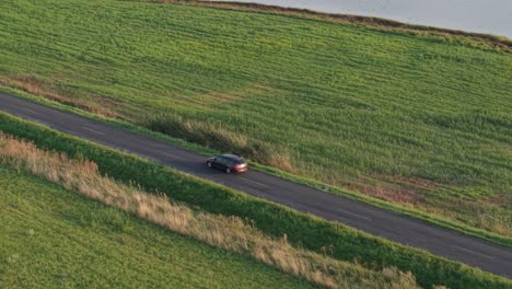 a brown car drives along a quiet asphalt road next to a lush green field and a calm lakeshore. the aerial perspective highlights the peaceful rural setting in warm evening light