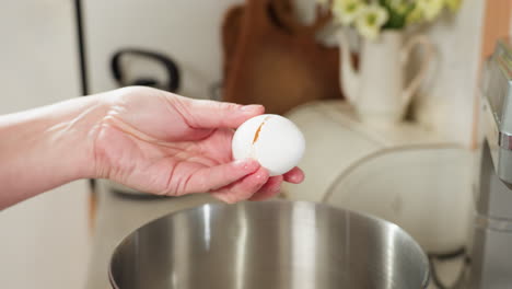 hand view of woman gently tapping egg with table knife above stainless bowl in kitchen, preparing to crack open shell with precision and care for cooking or baking