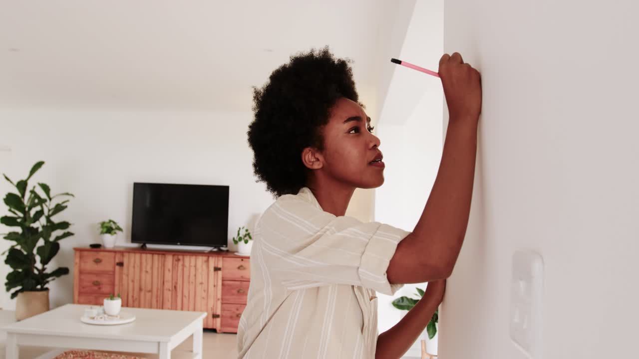 Woman mid twenties reaching for pink pencil on sofa, marking wall at home to hang frame
