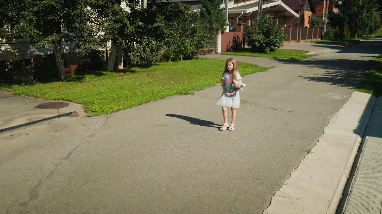Kid squinting under bright sunlight walks alone with hands locked together in calm suburban neighborhood, casting soft shadow on pavement with green grass and residential houses in background