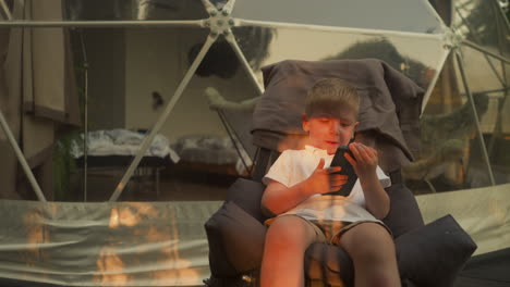 peaceful boy child plays with smartphone sitting in armchair on terrace of glamping house at sunset. relaxation alone with nature in quiet atmosphere slow motion