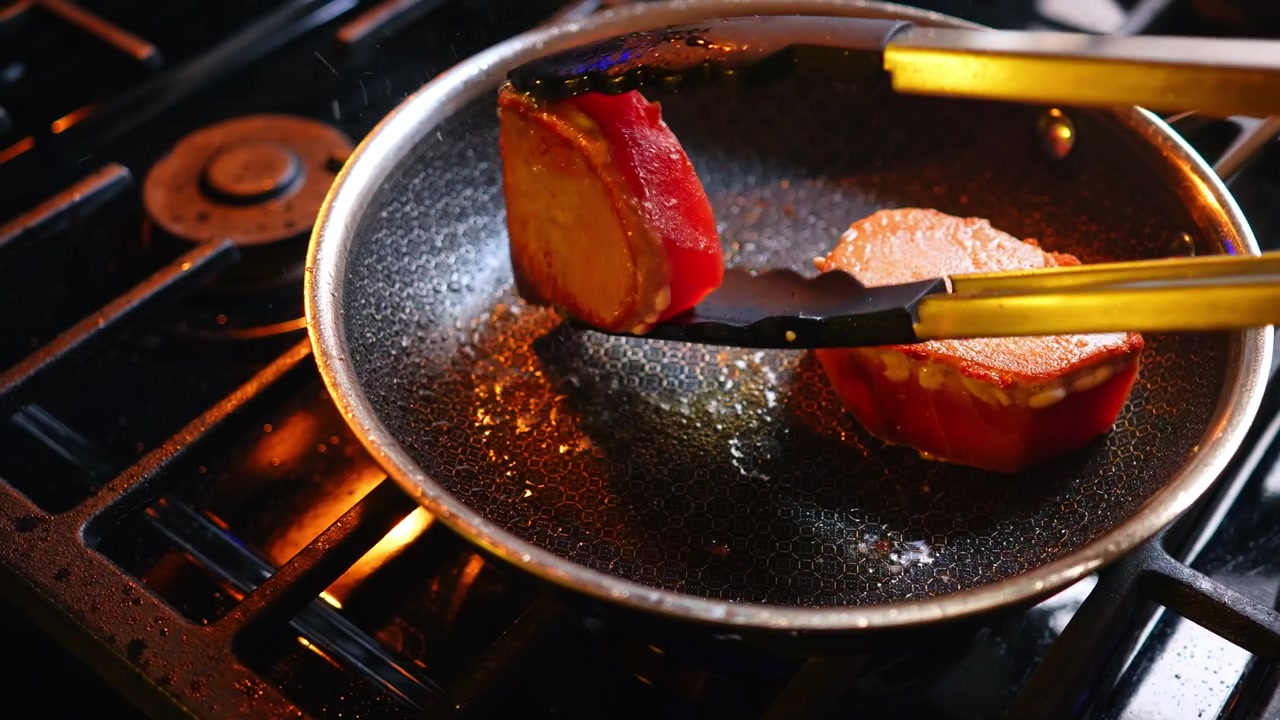 Metal tongs placing raw red tuna steak into frying pan. Close up of tongs holding fresh tuna fillet over stainless steel pan on gas stove burner with warm lighting.