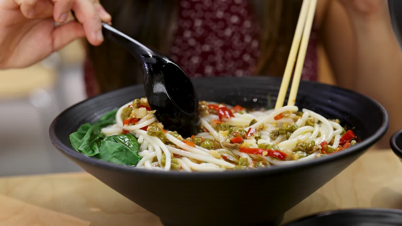 Close-up of woman stirring spicy noodle soup in black bowl, bright indoor lighting, shallow focus