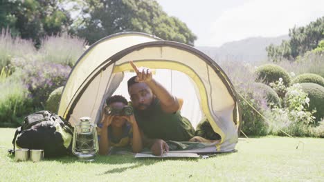 happy african american father and son sitting in tent and looking through binoculars, in slow motion