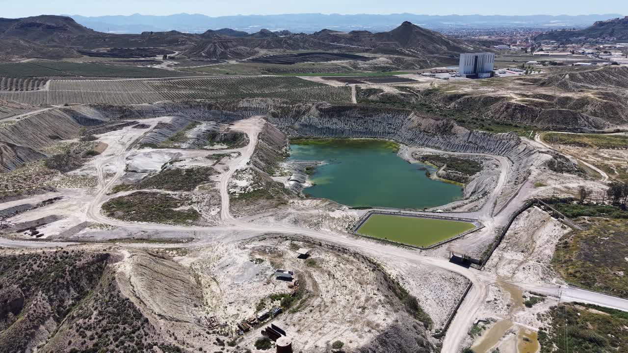 A tracking drone shot of a limestone quarry featuring a green water deposit and industrial infrastructure nestled in the arid hills of Murcia